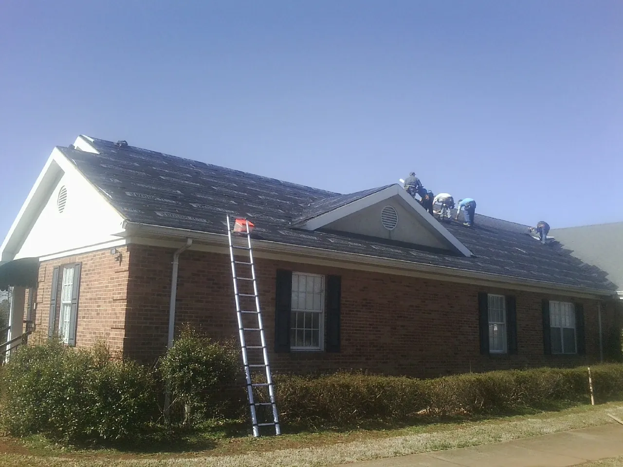 Crew tearing shingles off a brick ranch roof with a ladder set — Metal Roof Repair work by our crew in Keene